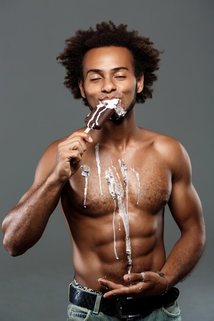 Hombre africano guapo joven comiendo helado, posando en gris