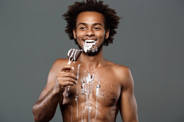 Hombre africano guapo joven comiendo helado, posando en gris