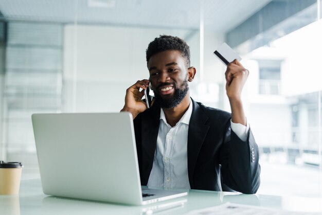 Hombre africano hablando por teléfono y leyendo el número de tarjeta de crédito mientras está sentado en la oficina