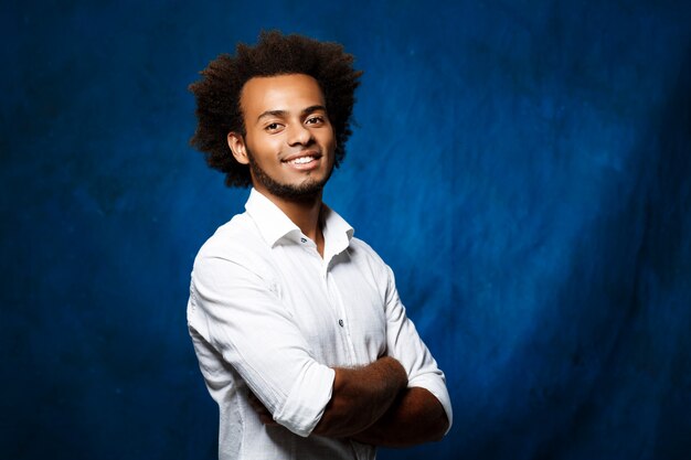 Hombre africano hermoso joven con los brazos cruzados sobre la pared azul.