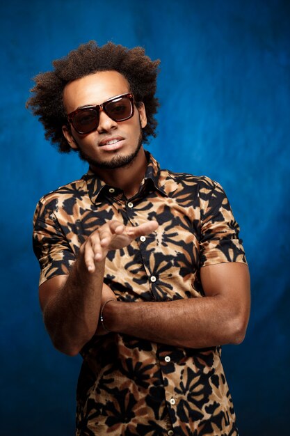 Hombre africano hermoso joven en las gafas de sol que presentan sobre la pared azul.