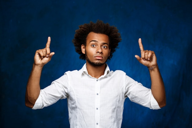 Hombre africano hermoso joven que destaca los dedos sobre la pared azul.