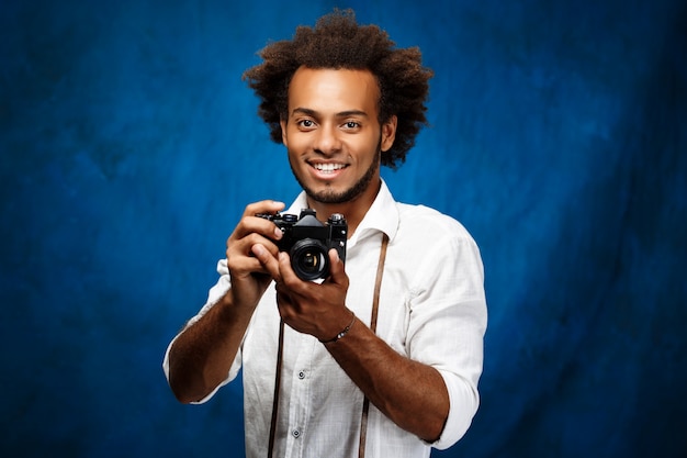 Hombre africano hermoso joven que sostiene la cámara vieja sobre la pared azul.