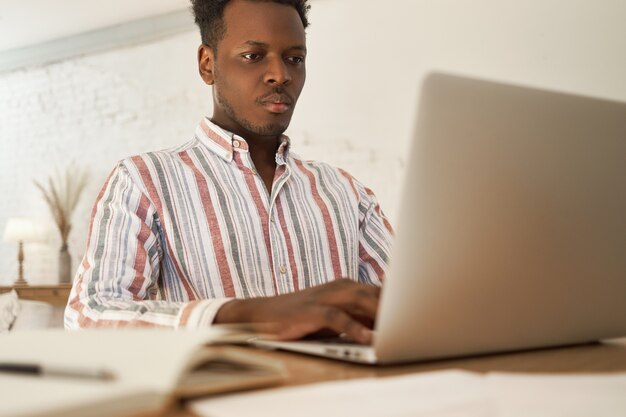 Hombre africano joven enfocado haciendo tareas independientes remotas en casa, buscando información usando wifi de alta velocidad.