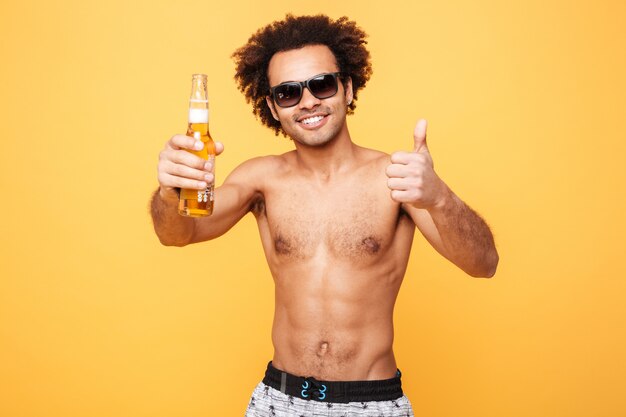 Hombre africano joven feliz bebiendo cerveza mostrando los pulgares para arriba.