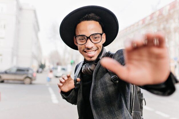 Hombre africano joven inspirado con sonrisa sincera posando sobre fondo borroso de la ciudad en la mañana. Chico guapo mulato pasar el día de otoño al aire libre.