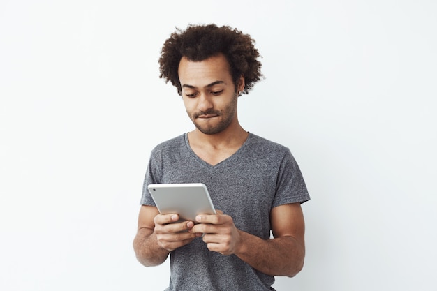 Hombre africano joven interesado y concentrado mirando tableta jugando un juego de plataformas y disfrutando de nuevos niveles sobre blanco filosófico.