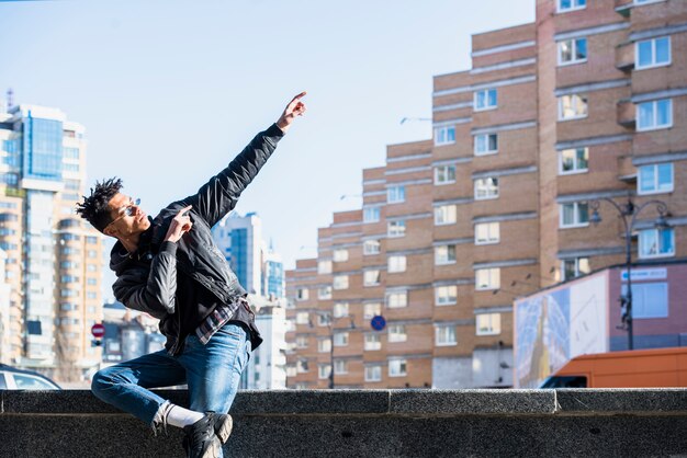 Hombre africano joven que se sienta en la pared que gesticula delante de edificios en la ciudad