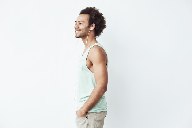 Hombre africano joven que sonríe escuchando transmitir música en auriculares con cable sobre la pared blanca. Locura habitual en sus veintes.