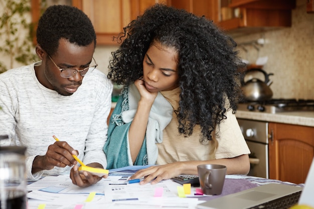 Hombre africano joven serio con gafas sosteniendo lápiz y papel, sentado en la mesa de la cocina con papeles y computadora portátil mientras calcula facturas y administra el presupuesto familiar junto con su esposa
