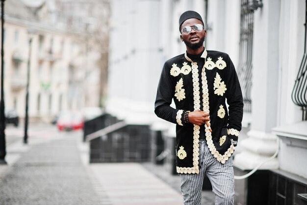 Hombre africano mega elegante en pose de chaqueta tradicional Chico negro de moda con sombrero y gafas de sol con cigarro en la mano