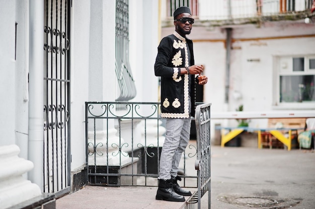 Hombre africano mega elegante en pose de chaqueta tradicional Chico negro de moda con sombrero y gafas de sol con cigarro en la mano