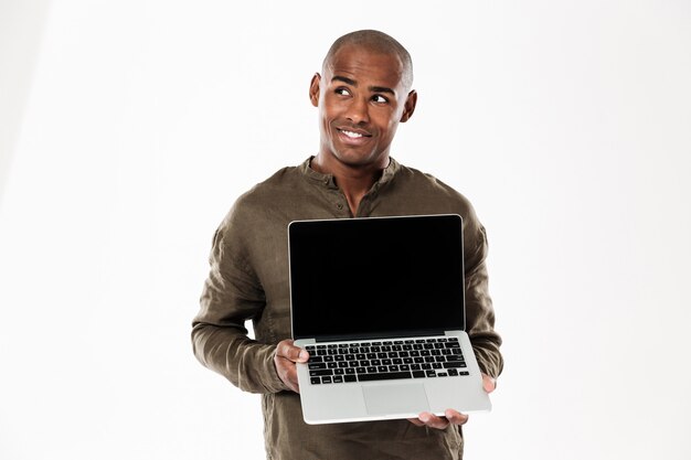 Hombre africano pensativo sonriente que muestra la pantalla de la computadora portátil en blanco y mirando hacia arriba