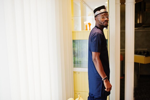 Hombre africano rico con ropa tradicional elegante y sombrero con gafas posadas en la habitación