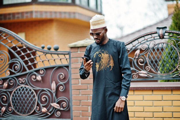 Hombre africano rico con ropa tradicional elegante y sombrero posado al aire libre en su mansión hablando por teléfono móvil