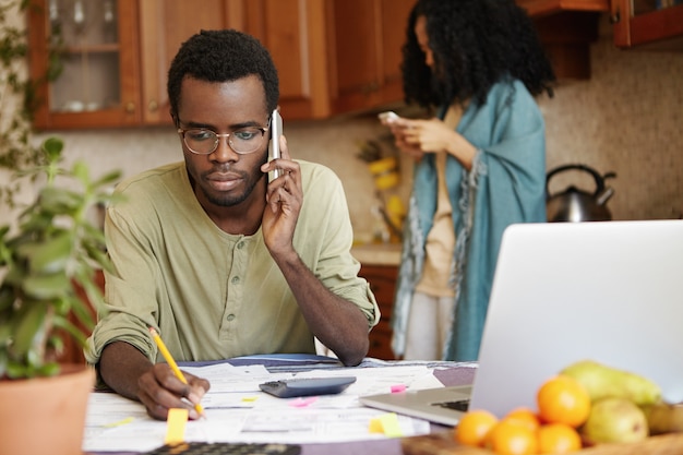 Hombre africano serio que tiene una conversación telefónica con el banco pidiendo extender el plazo del préstamo para pagar la hipoteca, sosteniendo un lápiz en la otra mano, tomando notas en documentos, acostado en la mesa frente a él