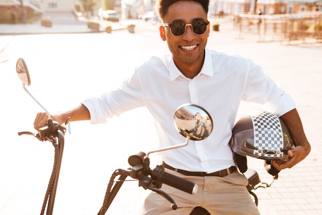 Hombre africano sonriente sentado en moto moderna al aire libre y mirando a la cámara