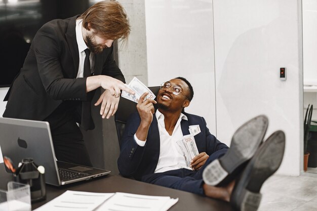 Hombre africano con traje negro. Chico sentado a la mesa. Hombre de negocios con dinero. Socios internacionales.