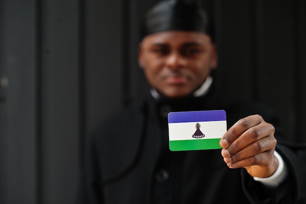 El hombre africano usa durag negro y sostiene la bandera de Lesotho en un fondo oscuro aislado a mano