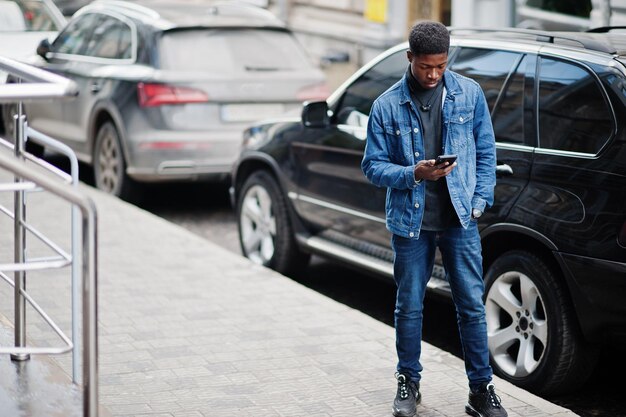 Hombre africano vestido con chaqueta de jeans posado al aire libre de pie contra un auto negro con teléfono móvil a mano