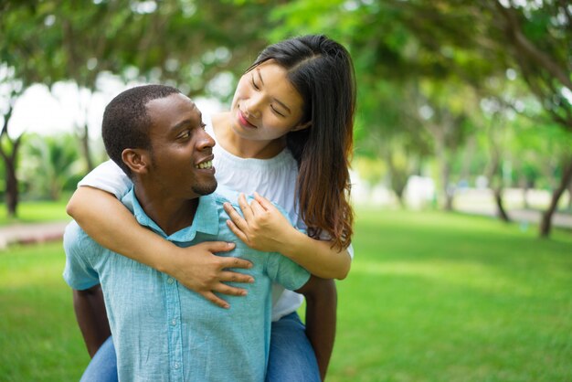 Hombre afroamericano alegre que lleva a cuestas asiático de la mujer.