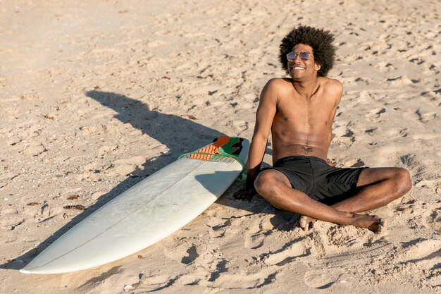 Hombre afroamericano alegre sentado en la arena con tabla de surf