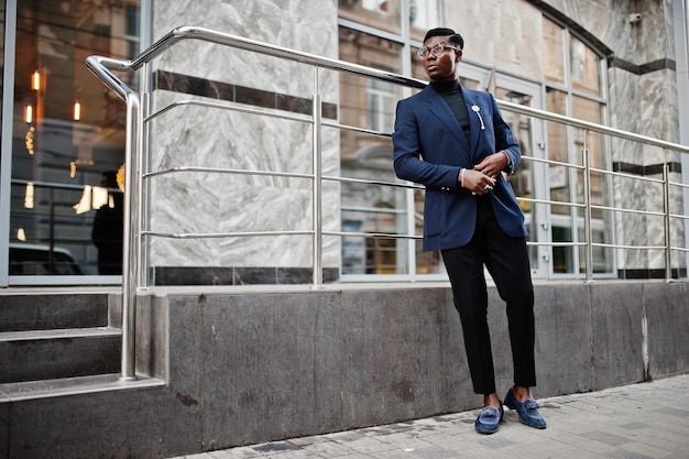 Foto gratuita un hombre afroamericano de aspecto asombroso usa una chaqueta azul con broche de cuello alto negro y gafas posadas en la calle chico negro de moda