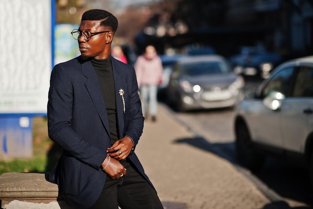 Un hombre afroamericano de aspecto asombroso usa una chaqueta azul con broche de cuello alto negro y gafas posadas en la calle Chico negro de moda