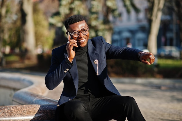 Un hombre afroamericano de aspecto asombroso usa una chaqueta azul con un broche de cuello alto negro y gafas posadas en la calle Un hombre negro de moda habla por teléfono y muestra algo