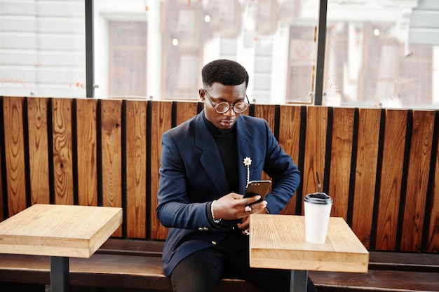 Un hombre afroamericano de aspecto asombroso usa una chaqueta azul con un broche de cuello alto negro y gafas posadas en la calle Un hombre negro de moda con una taza de café mirando su teléfono