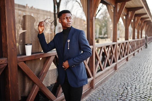 Un hombre afroamericano de aspecto asombroso usa una chaqueta azul con un broche de cuello alto negro y gafas posadas en la calle Un hombre negro de moda con una taza de café