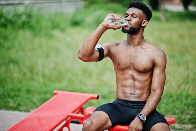 Hombre afroamericano atleta sexy deporte torso desnudo hombre con estuche de brazo deportivo para teléfono móvil haciendo ejercicios en los simuladores de entrenamiento de la calle y bebiendo agua de la botella