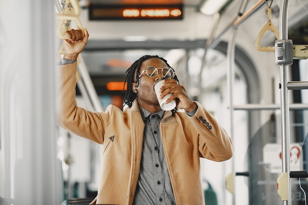 Hombre afroamericano en el autobús de la ciudad. Chico con un abrigo marrón. Hombre con café.