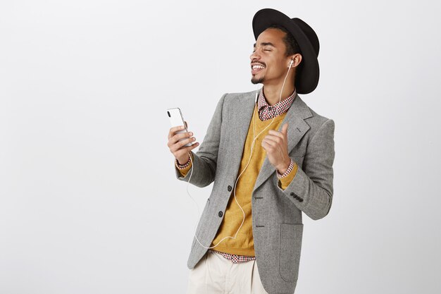 Hombre afroamericano bailando sin preocupaciones escuchando música en auriculares, sonriendo y sosteniendo el teléfono inteligente