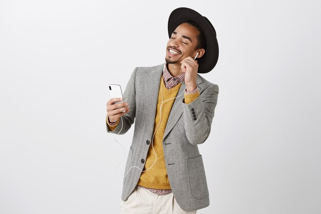 Hombre afroamericano bailando sin preocupaciones escuchando música en auriculares, sonriendo y sosteniendo el teléfono inteligente