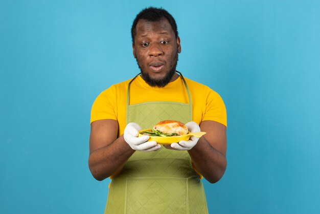 Hombre afroamericano con barba, delantal con un plato amarillo de sándwich en las manos, está parado sobre la pared azul.