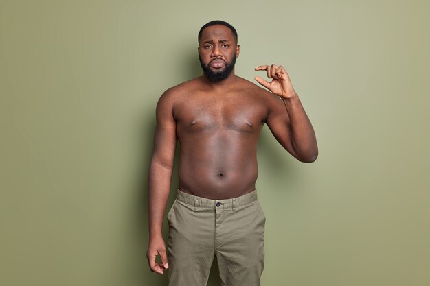 Hombre afroamericano barbudo disgustado posa con el torso desnudo muestra un gesto de cosita diminuta muestra poses de objeto muy pequeño contra la pared verde oscuro