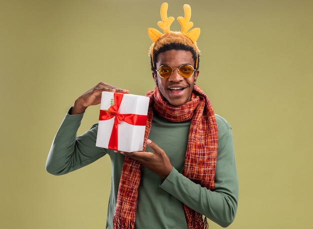 Hombre afroamericano con borde divertido con cuernos de ciervo y bufanda alrededor del cuello sosteniendo el regalo de Navidad mirando a la cámara con una sonrisa en la cara de pie sobre fondo verde