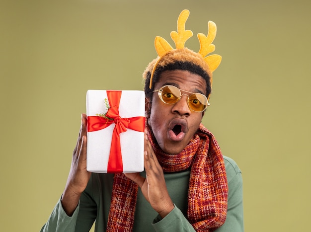 Hombre afroamericano con borde divertido con cuernos de ciervo y bufanda alrededor del cuello sosteniendo el regalo de Navidad mirando a la cámara sorprendido y asombrado de pie sobre fondo verde