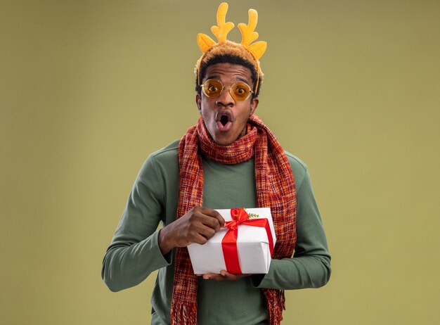 Hombre afroamericano con borde divertido con cuernos de ciervo y bufanda alrededor del cuello sosteniendo el regalo de Navidad mirando a la cámara sorprendido y asombrado de pie sobre fondo verde