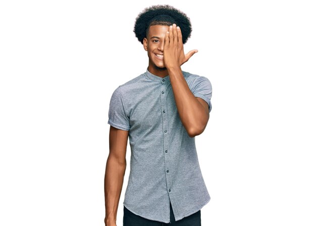 Foto gratuita hombre afroamericano con cabello afro vestido con ropa informal que cubre un ojo con una sonrisa segura en la cara y emoción sorpresa