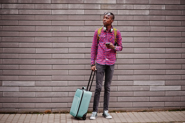 Hombre afroamericano en camisa a cuadros, gafas de sol y auriculares con maleta y mochila Viajero de hombre negro contra la pared con teléfono móvil