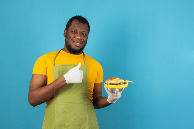 Hombre afroamericano comiendo comida sonriendo feliz y positivo, pulgar hacia arriba haciendo excelente y señal de aprobación, se encuentra sobre la pared azul claro.