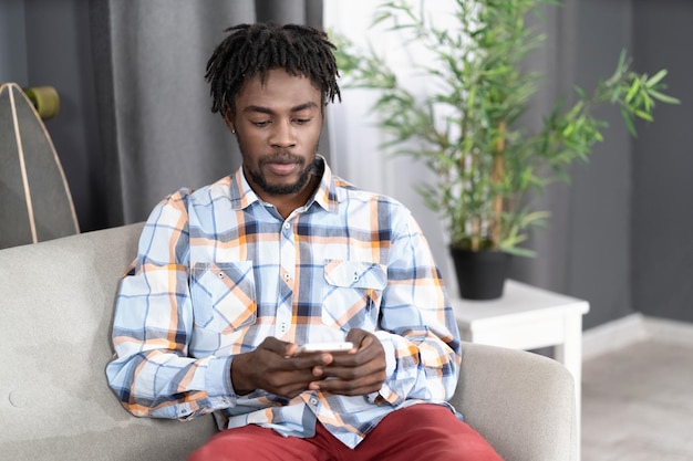 Un hombre afroamericano concentrado o interesado usa un teléfono inteligente revisando las redes sociales o actualizando información Retrato de un joven sosteniendo un teléfono inteligente sentado en el sofá Concepto de redes sociales