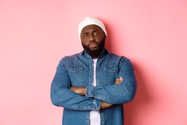 Hombre afroamericano decepcionado en gorro hipster, mirando a la cámara disgustado, brazos cruzados sobre el pecho, de pie sobre fondo rosa