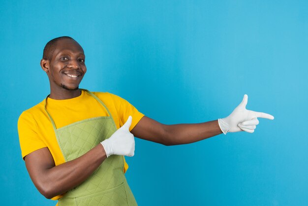 Hombre afroamericano en delantal verde sosteniendo dando pulgar hacia arriba en la pared azul
