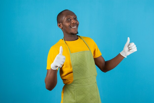 Hombre afroamericano en delantal verde sosteniendo dando pulgar hacia arriba en la pared azul