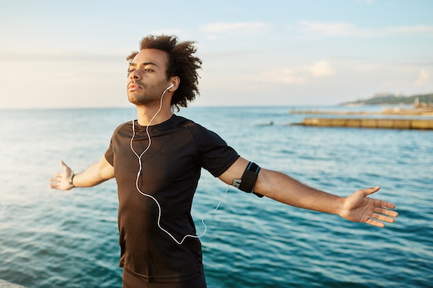 Hombre afroamericano deportivo estirando sus brazos antes de hacer ejercicio al aire libre. Atleta masculino delgado y fuerte con camiseta negra, manteniendo los brazos abiertos, respirando aire fresco del mar.