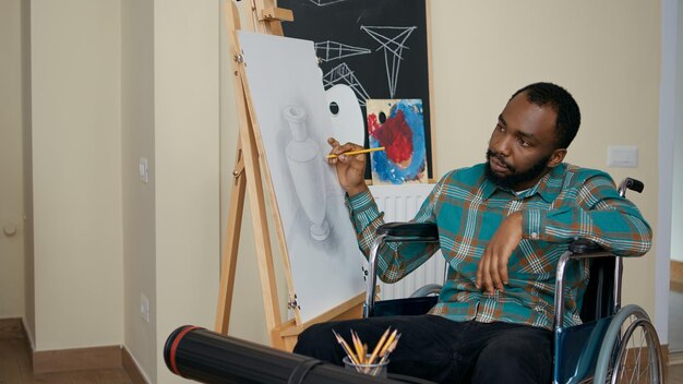 Hombre afroamericano con discapacidad usando lápiz para dibujar sobre lienzo, aprendiendo nuevas habilidades artísticas en clase de arte con el maestro. Joven estudiante en silla de ruedas dibujando una obra maestra en el taller.