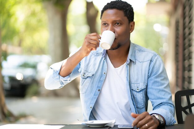 Hombre afroamericano disfrutando y bebiendo un café mientras está sentado en la cafetería al aire libre